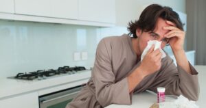 0_Man-sitting-at-table-with-tissue-blowing-nose-and-holding-head.jpg